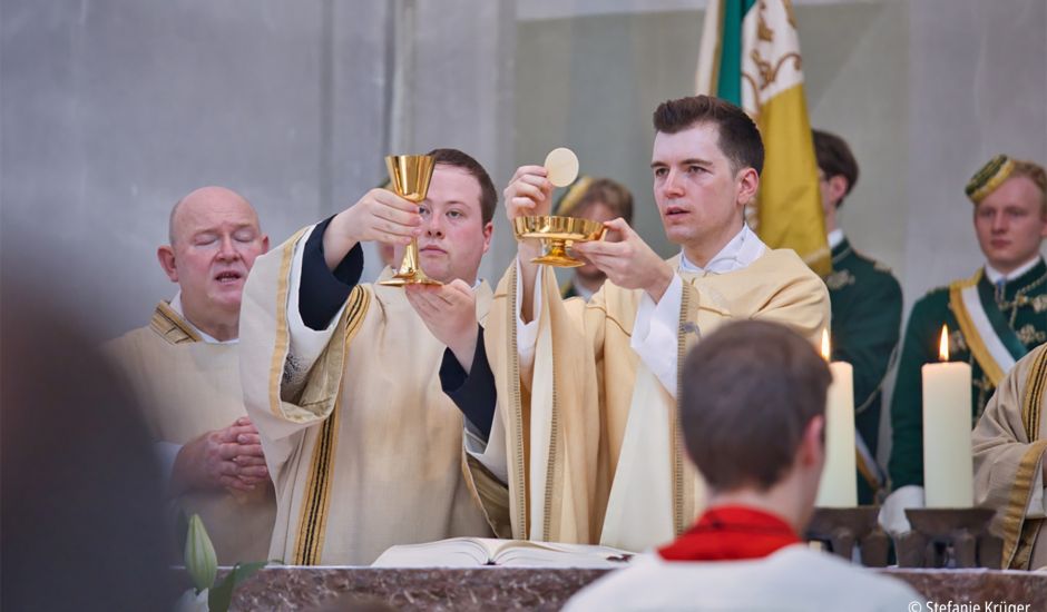 Zwei Priester stehen am Altar, einer hält einen Kelch, ein anderer eine Schale mit Hostien in die Höhe