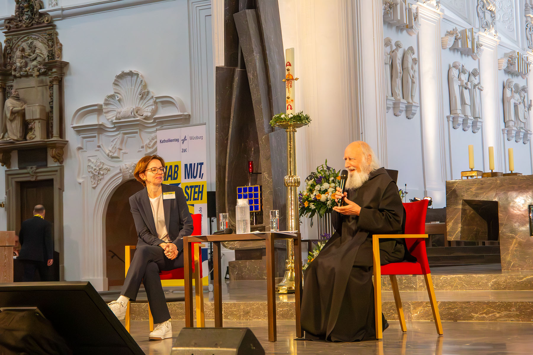 Frau und Mönch mit weißem Bart sitzen auf roten Stühlen in einer Kirche