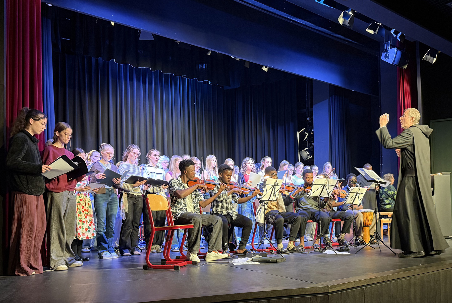 Große Gruppe an Schülerinnen und Schülern auf der Bühne, vorne Gastschüler aus Tansania mit Musikinstrumenten