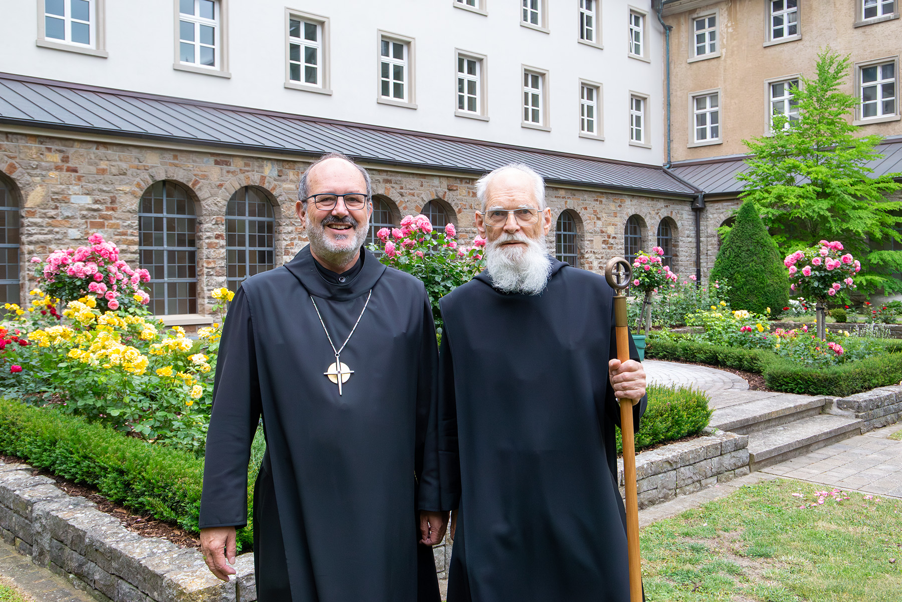Zwei Mönche mit schwarzen Gewand stehen vor Rosenbeeten