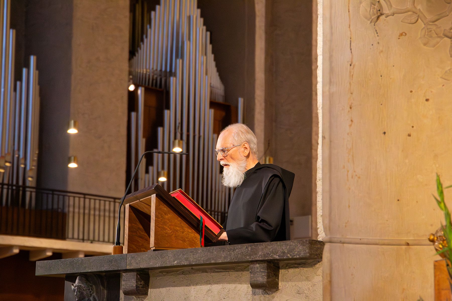 Mönch im Habit vor einem Lesepult, hinter ihm sieht man eine große Orgel