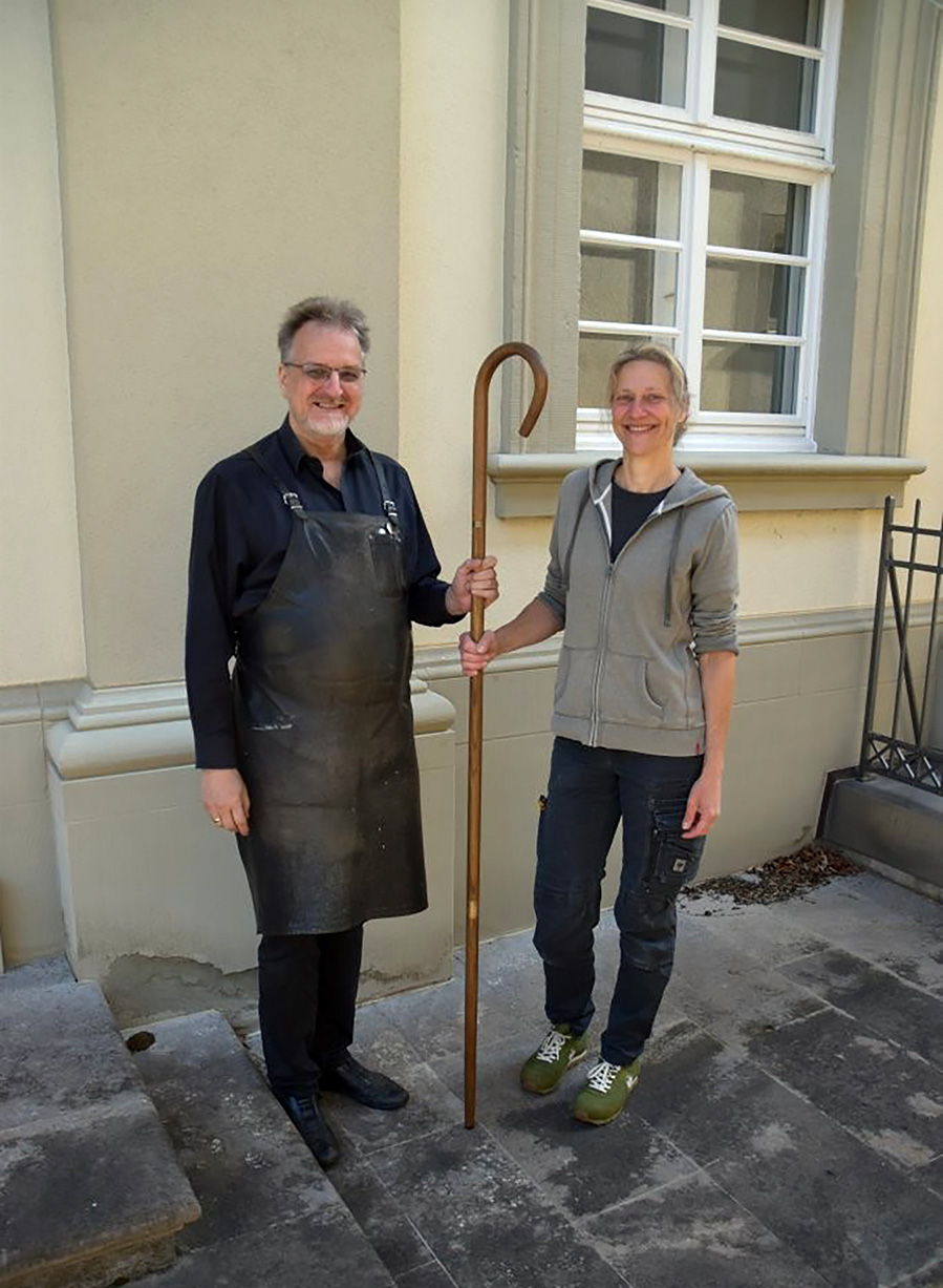 Ein Mann in Schürze und eine Frau halten gemeinsam den Bischofstab