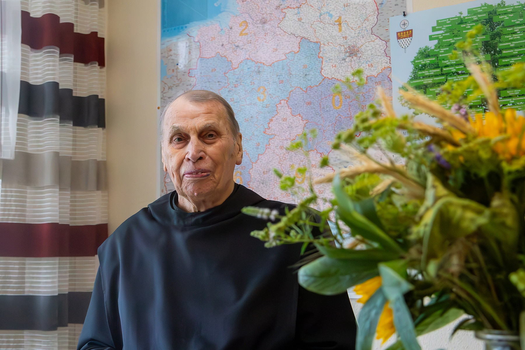 Älterer Mönch in einem Büro vor einer Landkarte, neben ihm ein Strauß Sonnenblumen