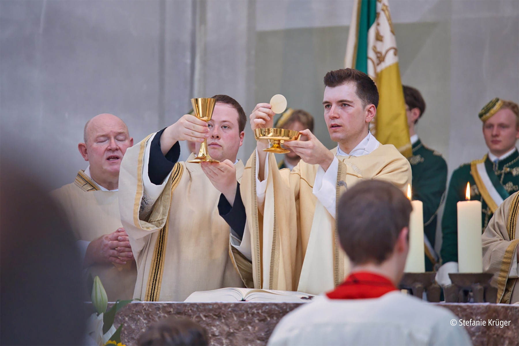 Zwei Priester stehen am Altar, einer hält einen Kelch, ein anderer eine Schale mit Hostien in die Höhe