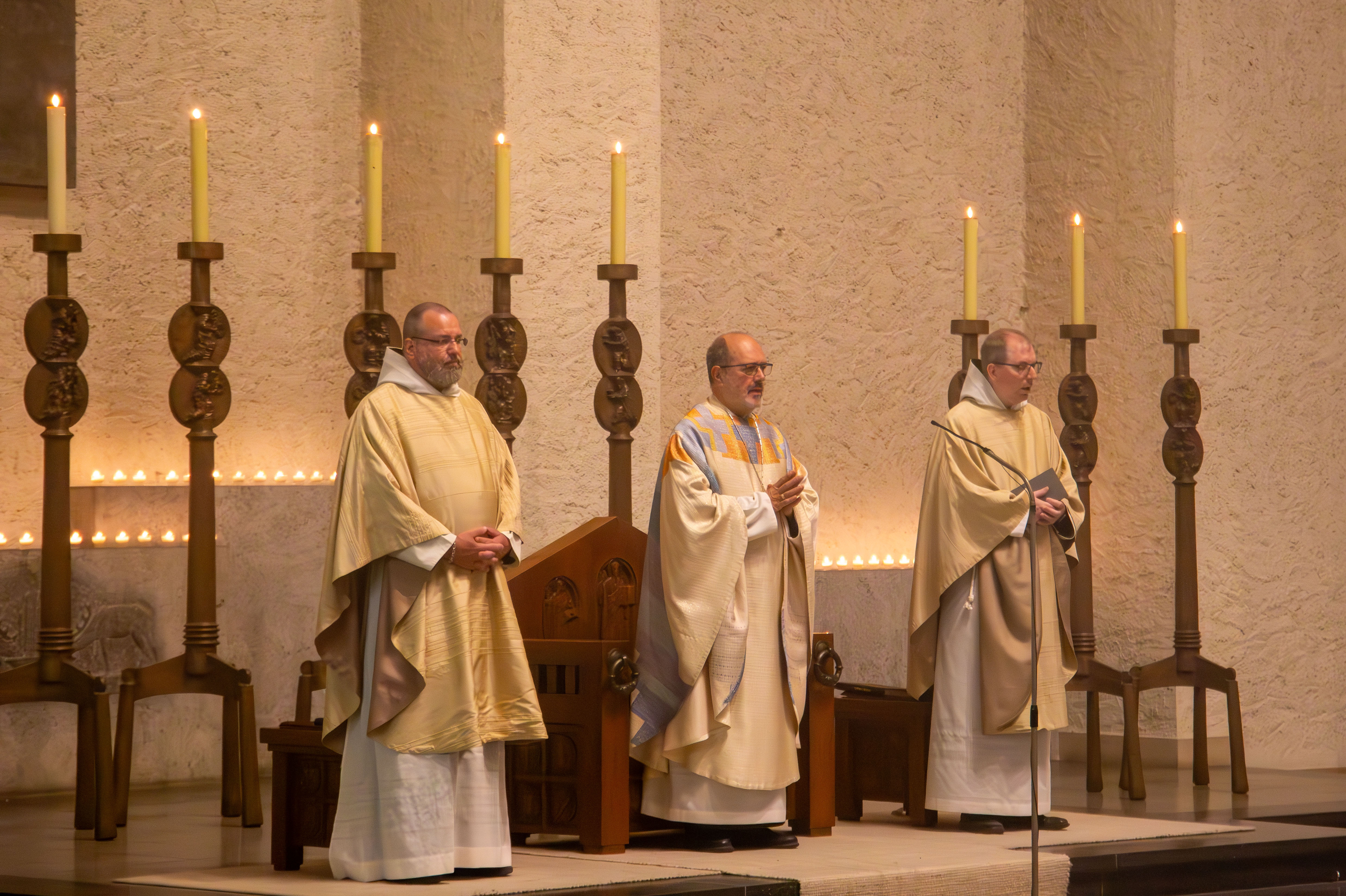 Drei Mönche in hellen Gewändern stehen mit betenden Händen in der Kirche