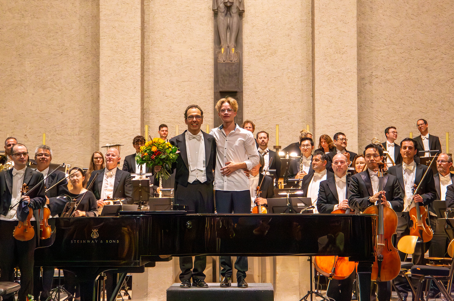 Dirigent mit Blumen und junger Mann stehen vor Orchester