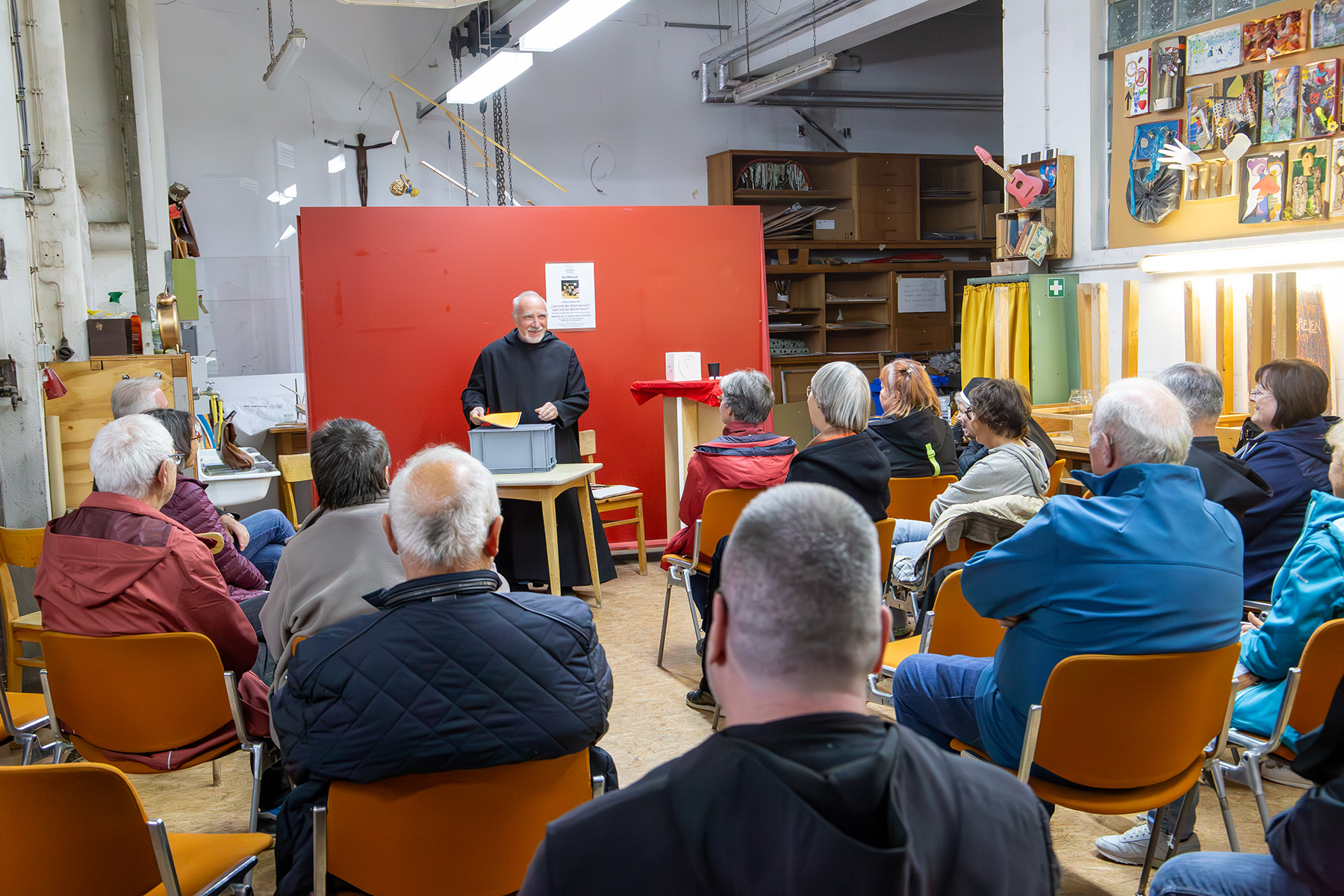 Mönch vor roter Wand redet mit dem Publikum