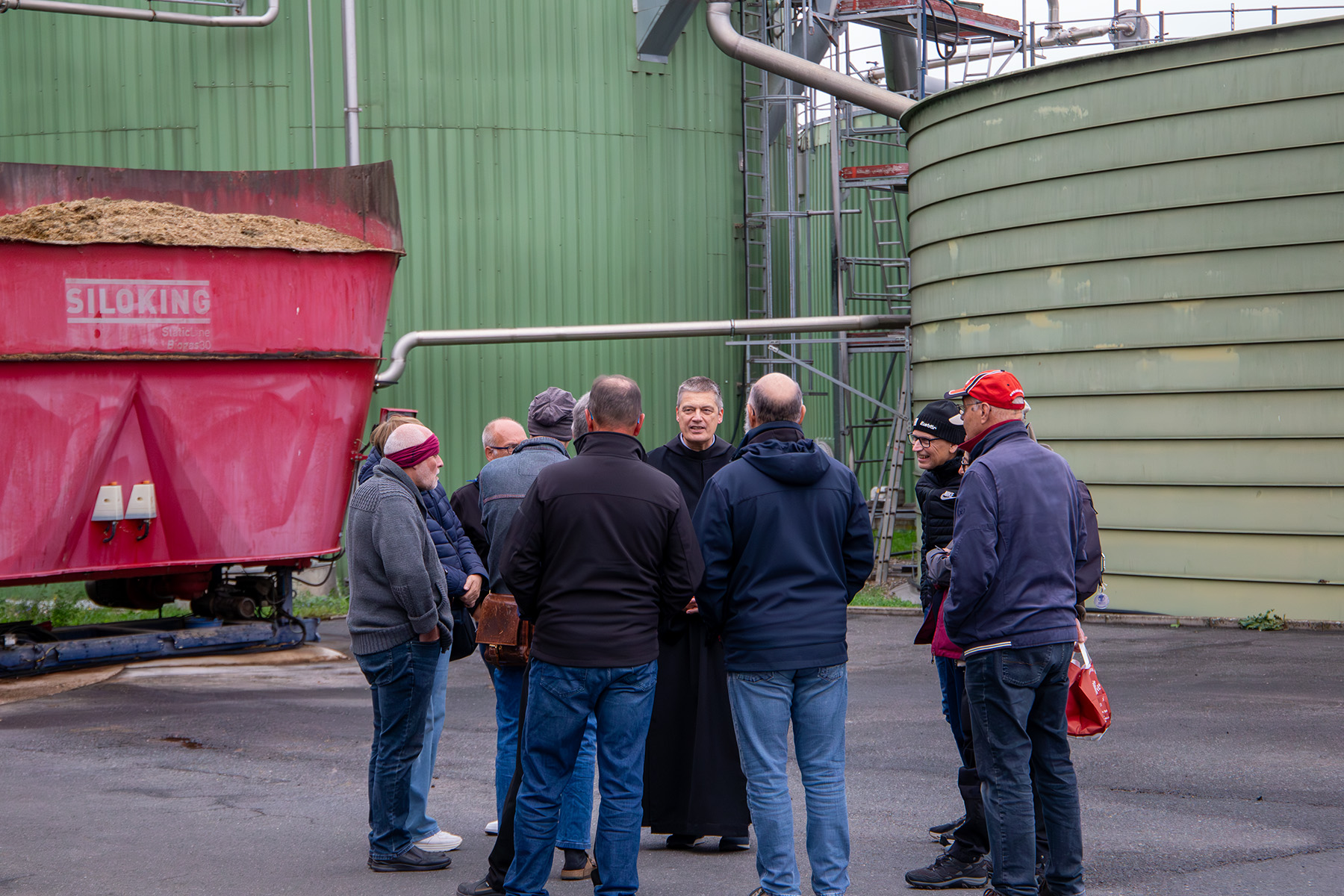 Menschen stehen um einen Mönch vor landwirtschaftlichen Anlagen