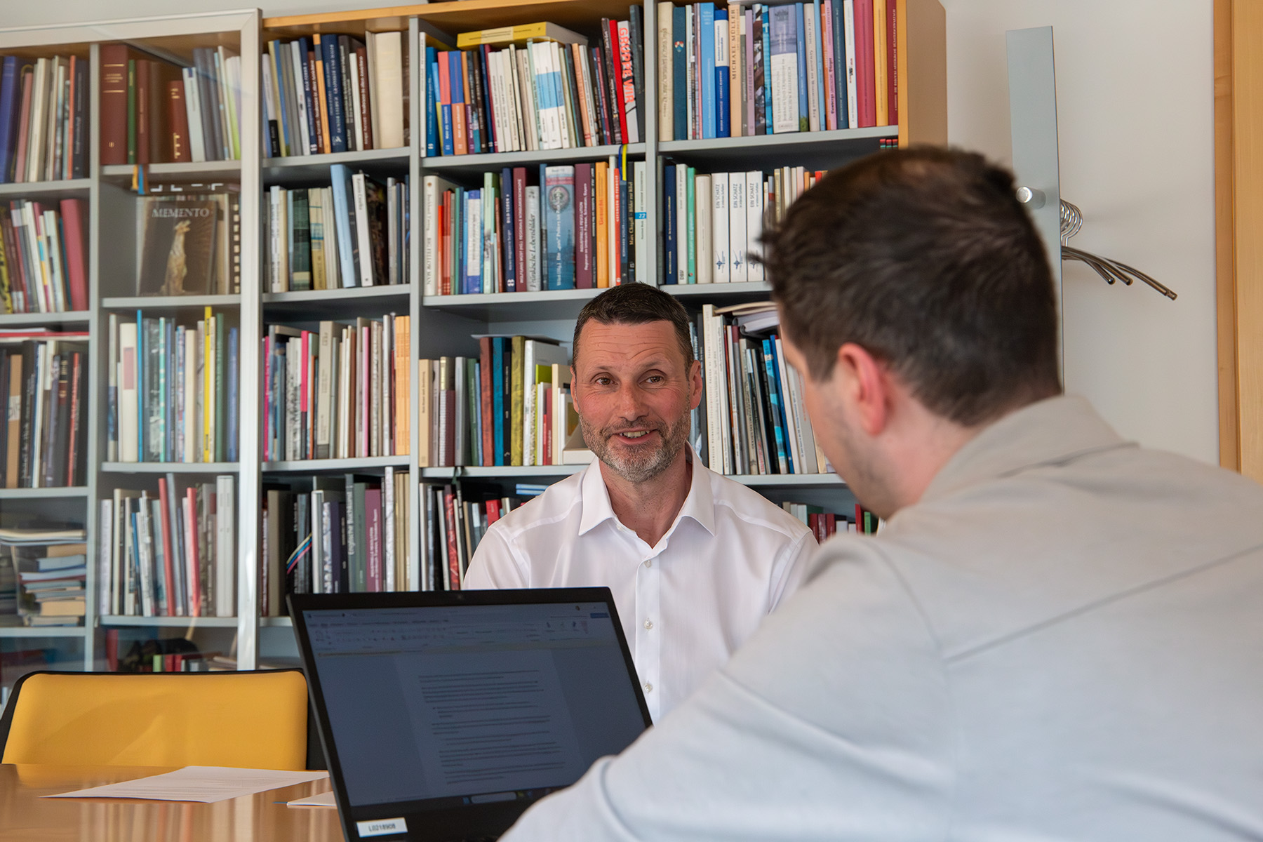 Zwei Männer unterhalten sich, im Hintergrund ein Buchregal, auf dem Tisch ein Laptop