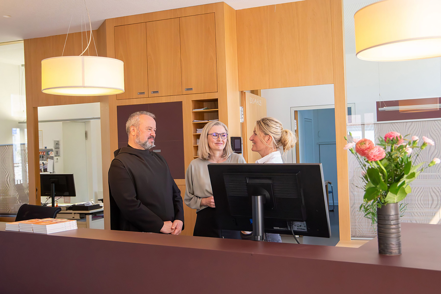 Mönch und zwei Frauen stehen an einer Rezeption und unterhalten sich
