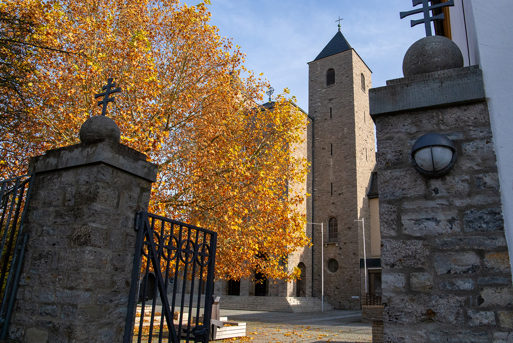 Blick durch ein offenes Tor auf einen Baum mit bunten Blättern sowie einer Kirche daneben