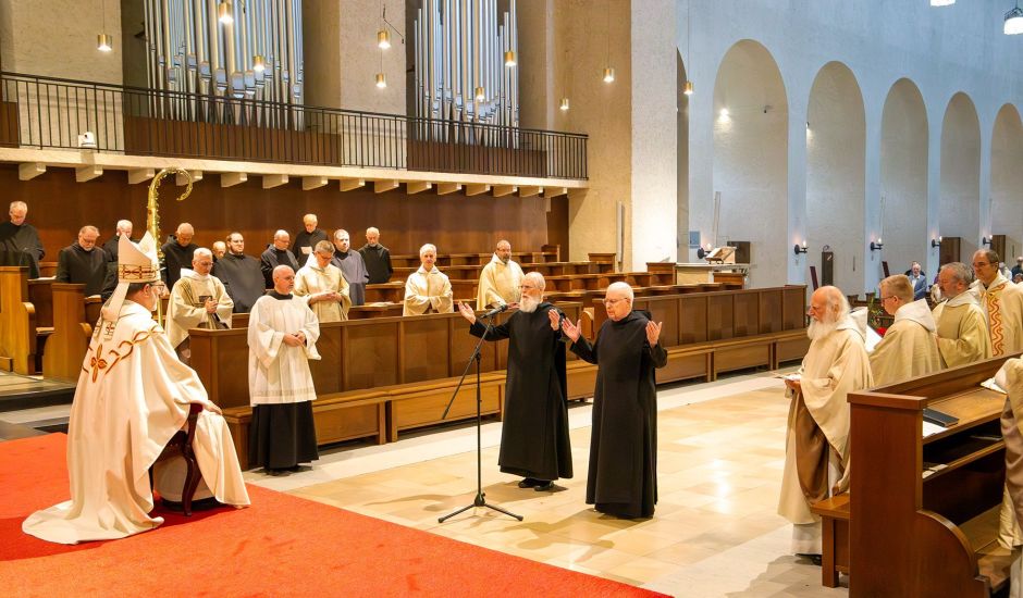 Zwei Mönche stehen mit erhobenen Händen in der Kirche vor einem Mann in weißem Gewandt und mit Abthut