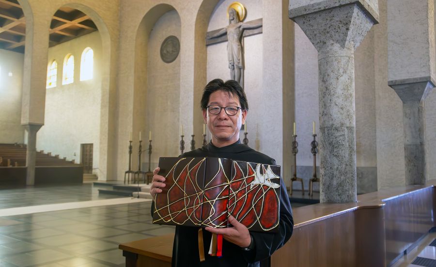 Mönch im schwarzen Gewand mit einem großen Buch in einer Kirche, im Hintergrund steht ein großes Kreuz