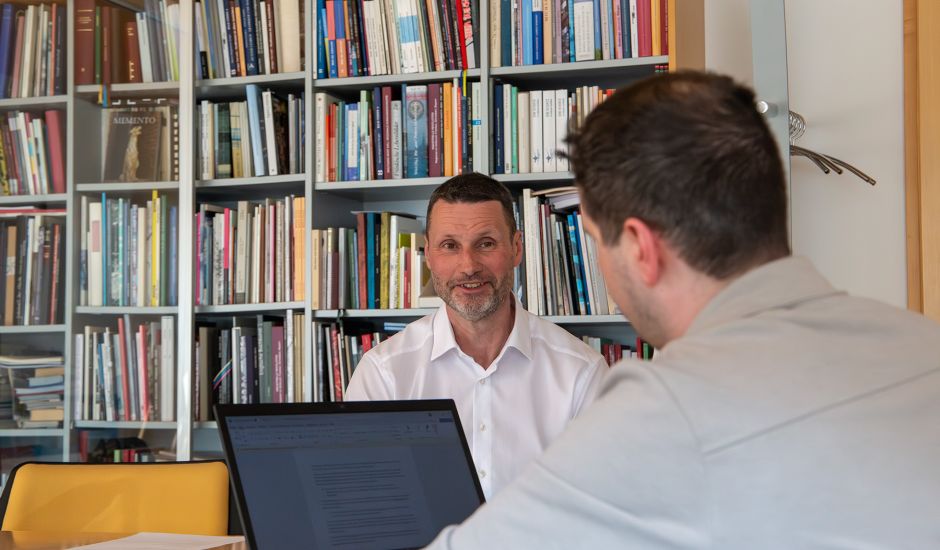 Zwei Männer unterhalten sich, im Hintergrund steht ein Buchregal, auf dem Tisch ist ein Laptop