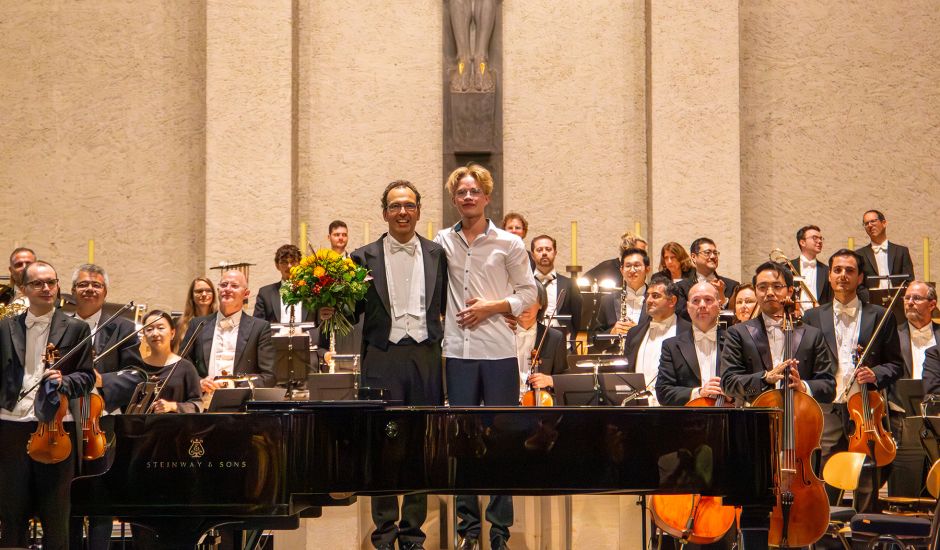 Mann im Frack und ein junger Mann stehen vor einem Orchester