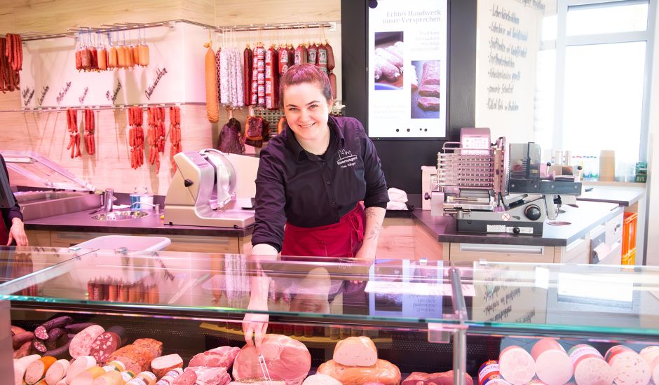 Junge Frau hinter einer Frischetheke sticht mit einer Gabel in eine Wurst