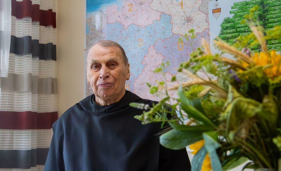 Älterer Mönch in einem Büro vor einer Landkarte, neben ihm ein Strauß Sonnenblumen