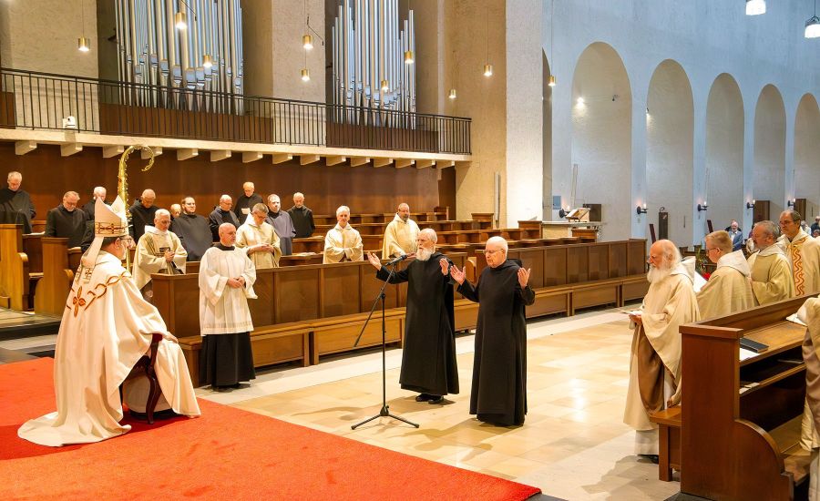 Zwei Mönche stehen mit erhobenen Händen in der Kirche vor einem Mann in weißem Gewandt und mit Abthut