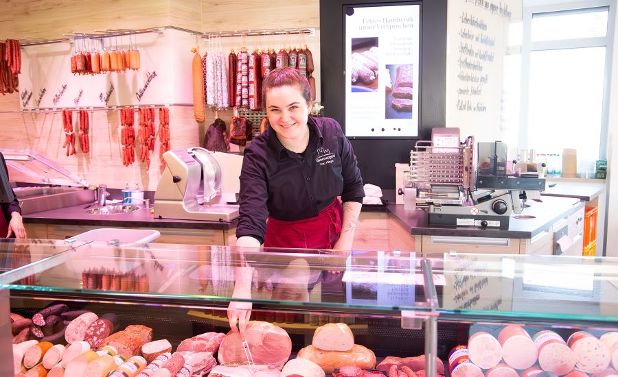 Junge Frau hinter einer Frischetheke sticht mit einer Gabel in eine Wurst