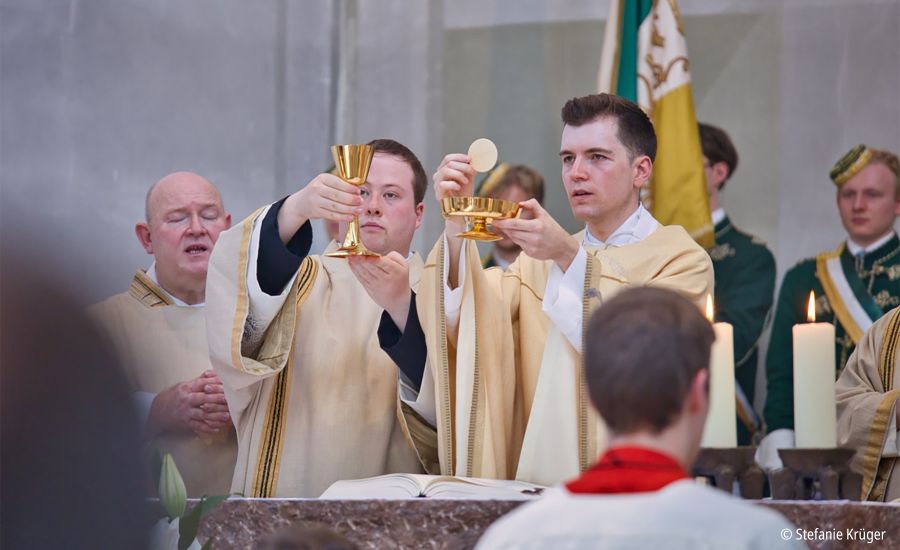 Zwei Priester stehen am Altar, einer hält einen Kelch, ein anderer eine Schale mit Hostien in die Höhe