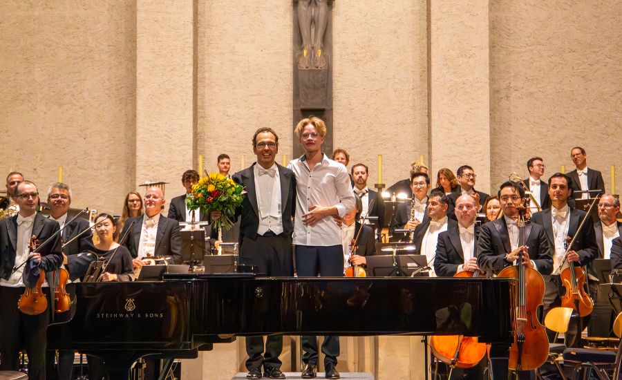 Mann im Frack und ein junger Mann stehen vor einem Orchester