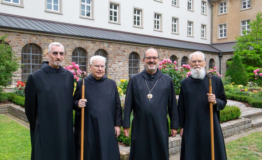 Vier Mönche im schwarzen Habit stehen in einem Garten, zwei halten einen braunen Stab in der Hand