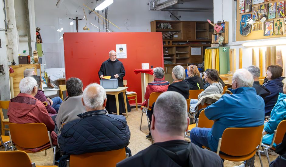 Mönch steht vor einer  roten Rückwand und spricht vor einem Publikum
