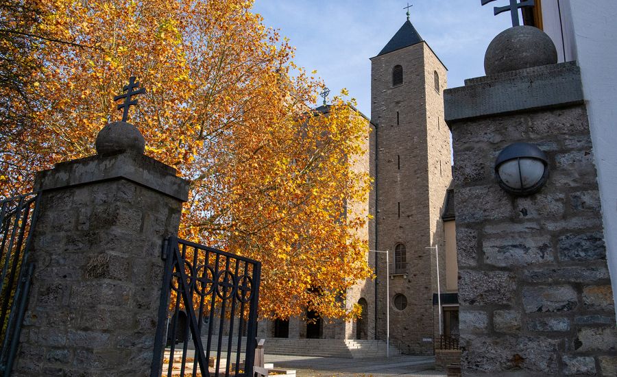 Blick durch ein offenes Tor auf einen Baum mit bunten Blättern sowie einer Kirche daneben