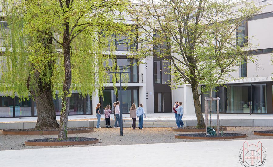 Blick auf ein Schulgebäude, vor dem Jugendliche sowie mehrere Bäume stehen